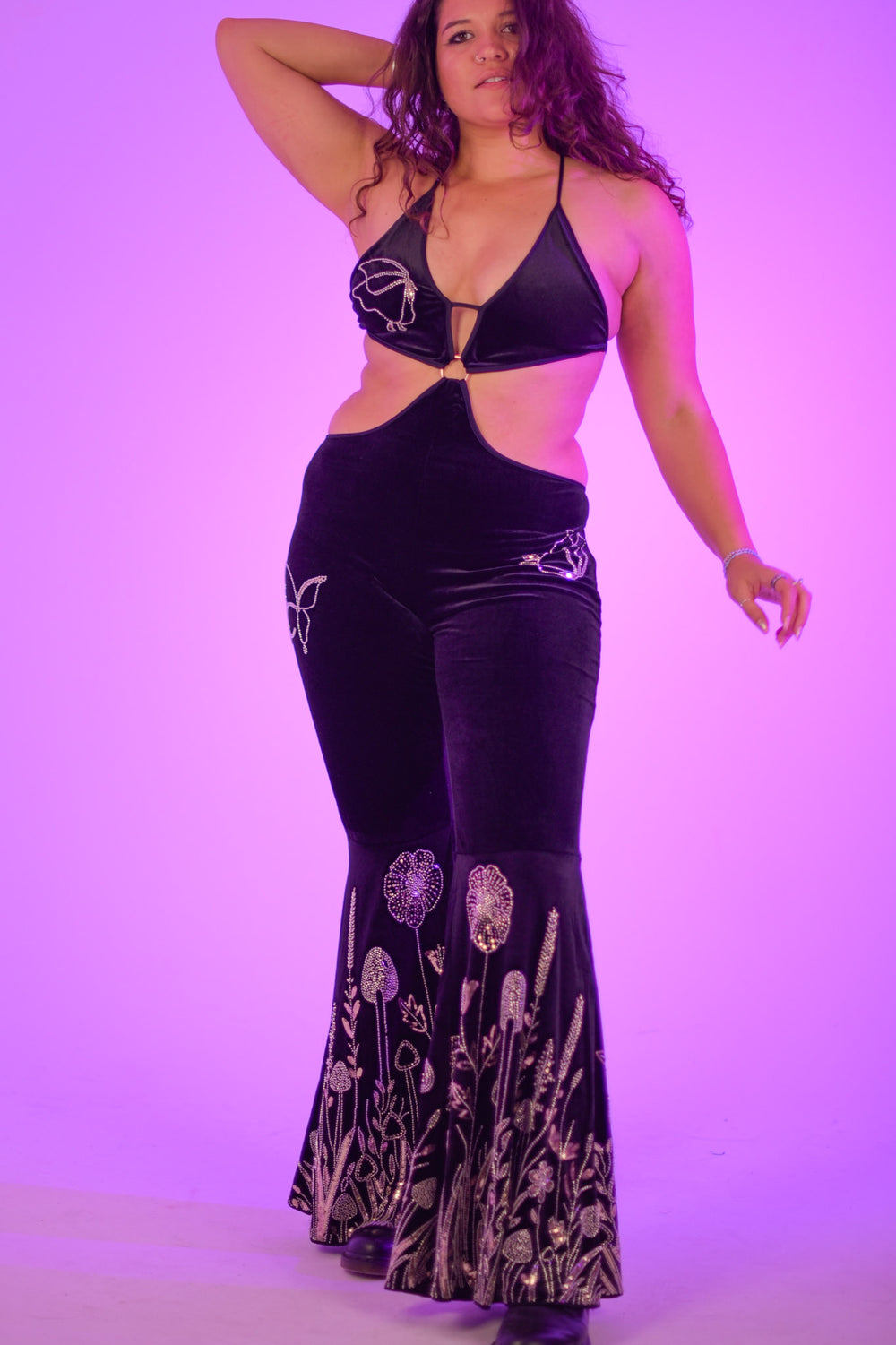 Plus-size model wearing The Fairy Garden Catsuit, showcasing velvet fabric and glittering botanical designs on flared legs. Festival-ready statement piece.