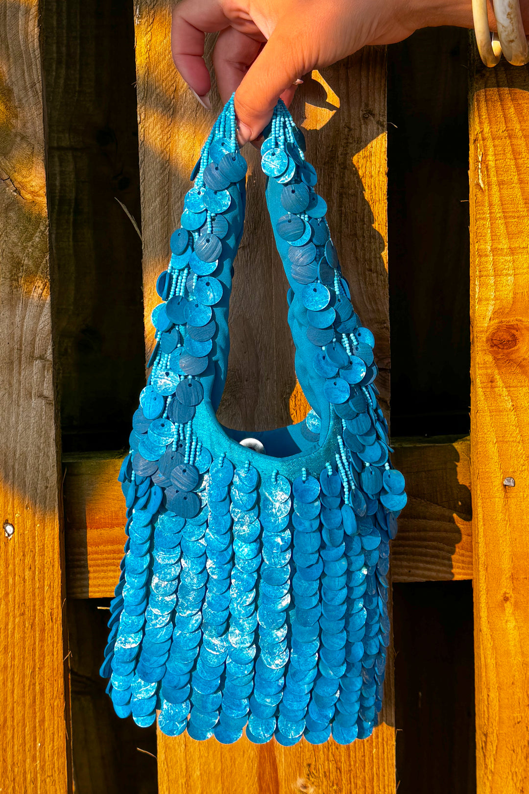 Oceana Shell Blue Bag with shimmering blue sequins, perfect for rave and festival outfits. Handheld against a wooden fence in sunlight.